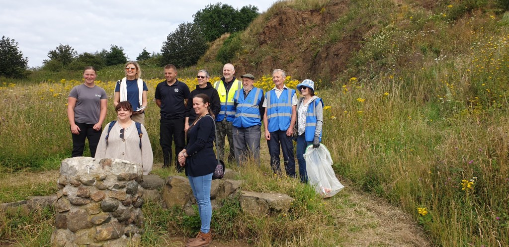 Image shows a group of people behind a cairn stone on Portway Hill with backdrop of grass and a rock face.